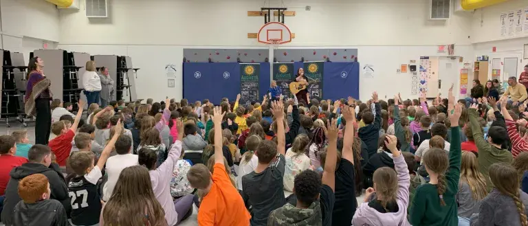 Children sitting in a gym raising their hands excitedly