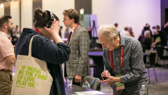 a person listening to a composer's work through headphones as the composer in front of her clicks play on his device