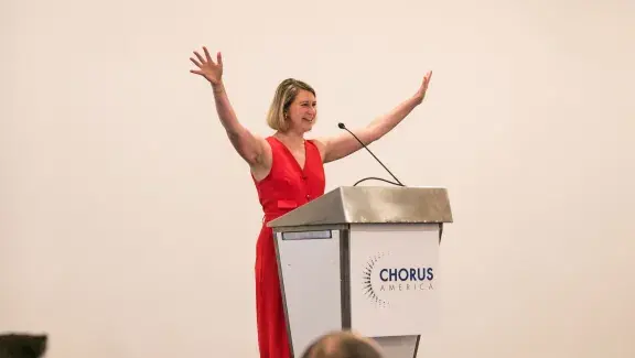 Emily Isaacson presenting in front of room and waving her hands in the air with a podium in front of her