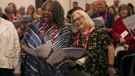 Two women standing next to each other and holding sheet music while singing and smiling